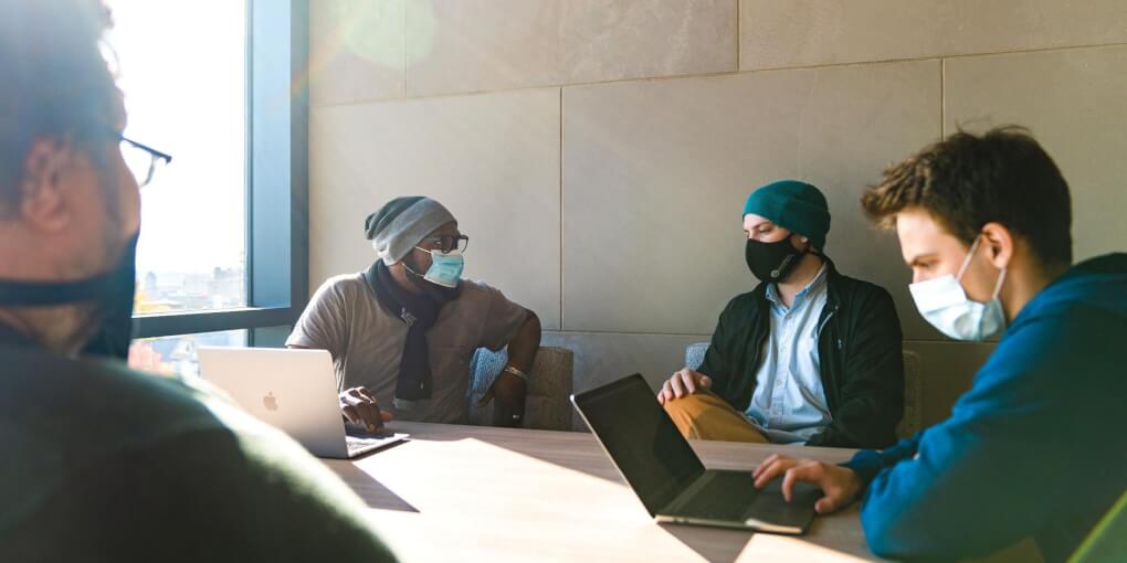 A group of four students sits around a table in the library wearing face masks and using their laptops